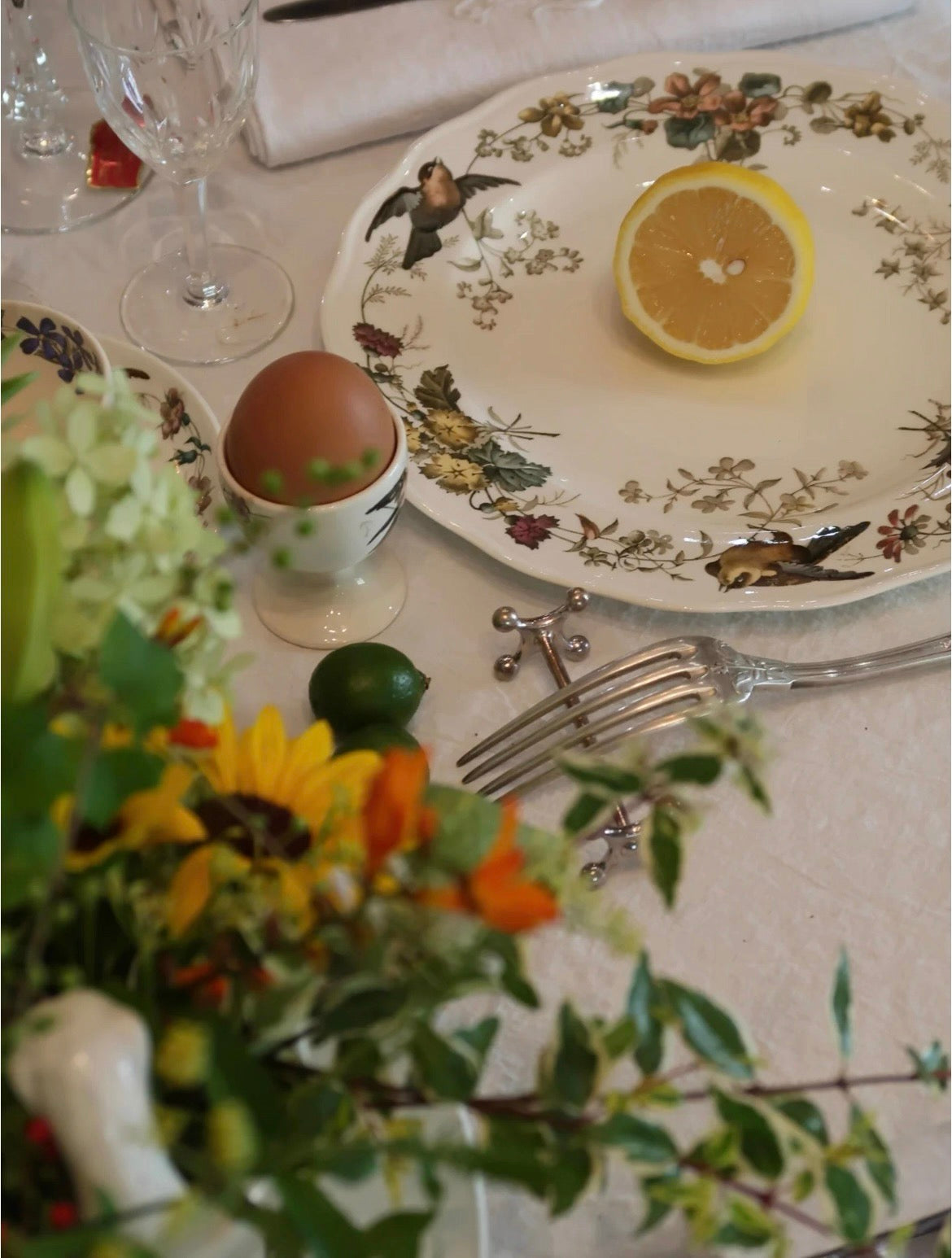 Table setting with floral arrangement, lemon, egg, and silverware on a decorative plate.