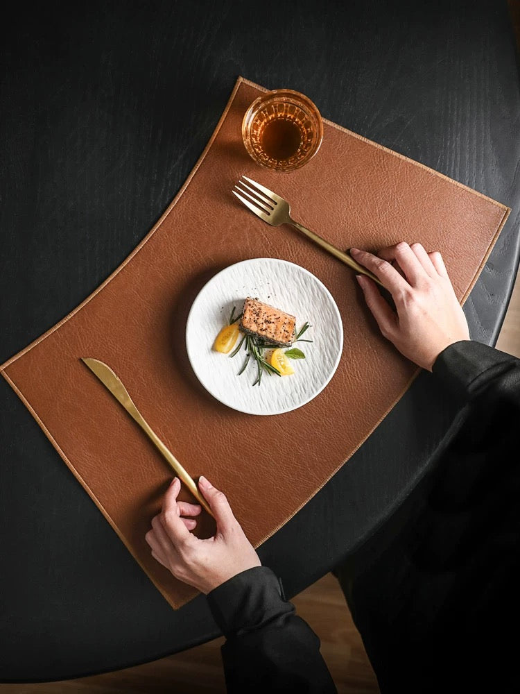 Person setting a table with a plate of food, fork, knife, and glass on a brown placemat.