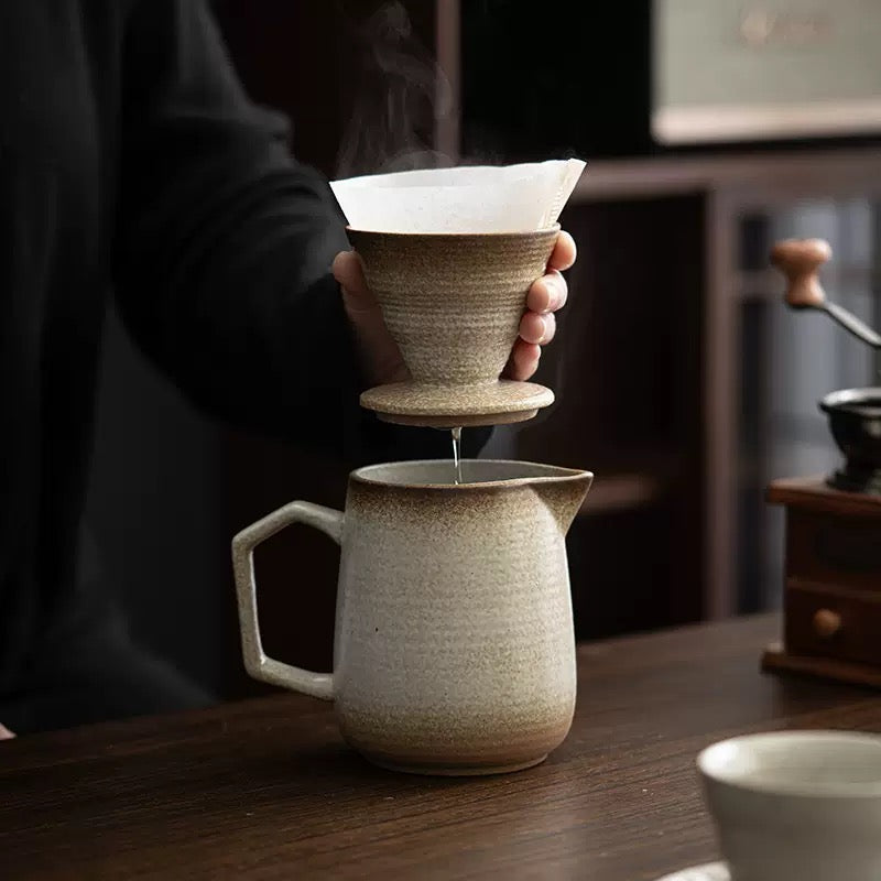 Person pouring coffee from a ceramic pitcher into a filter over a cup on a wooden table.