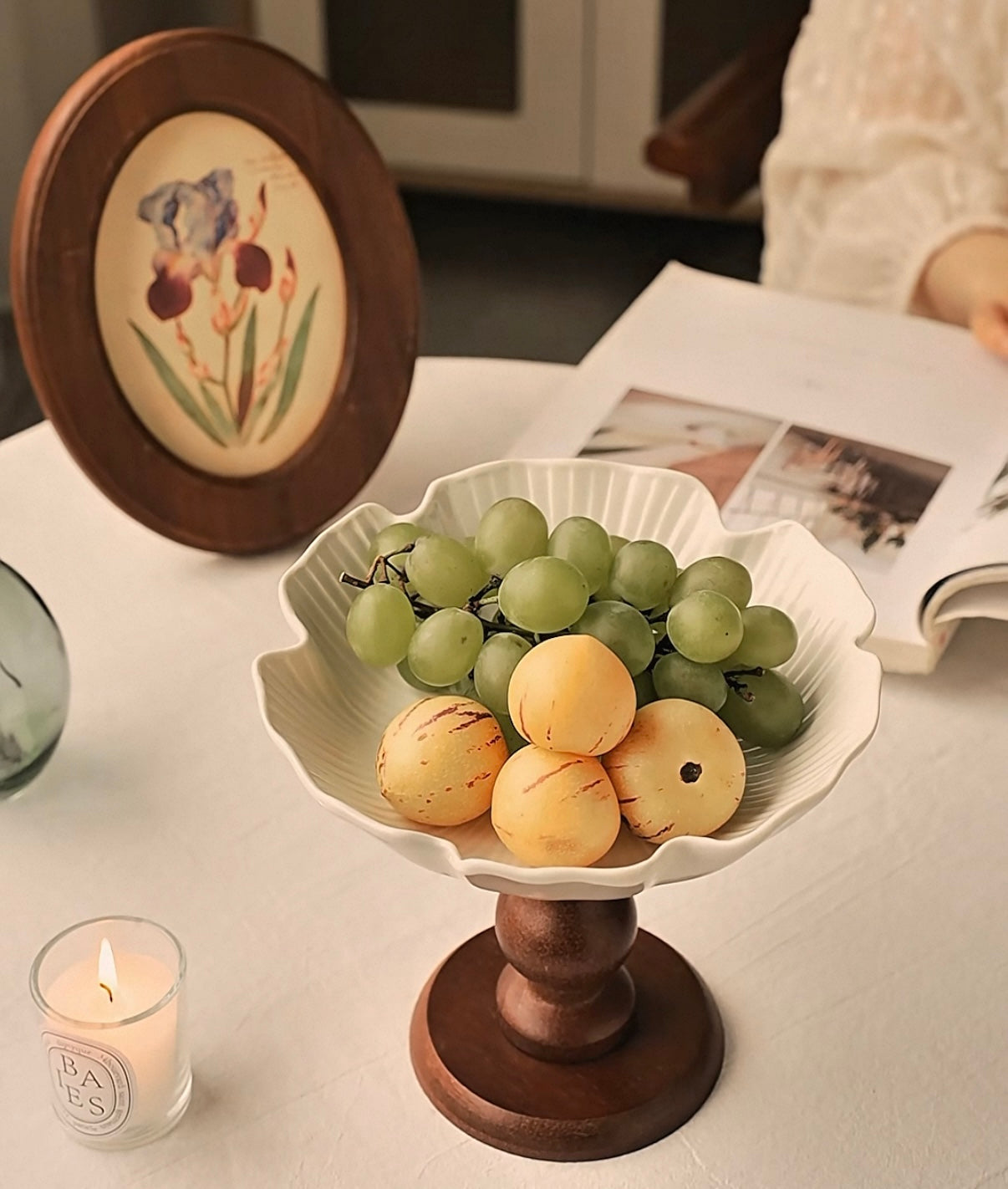 Decorative fruit bowl with grapes and pears on a wooden stand, candle, and book in the background.