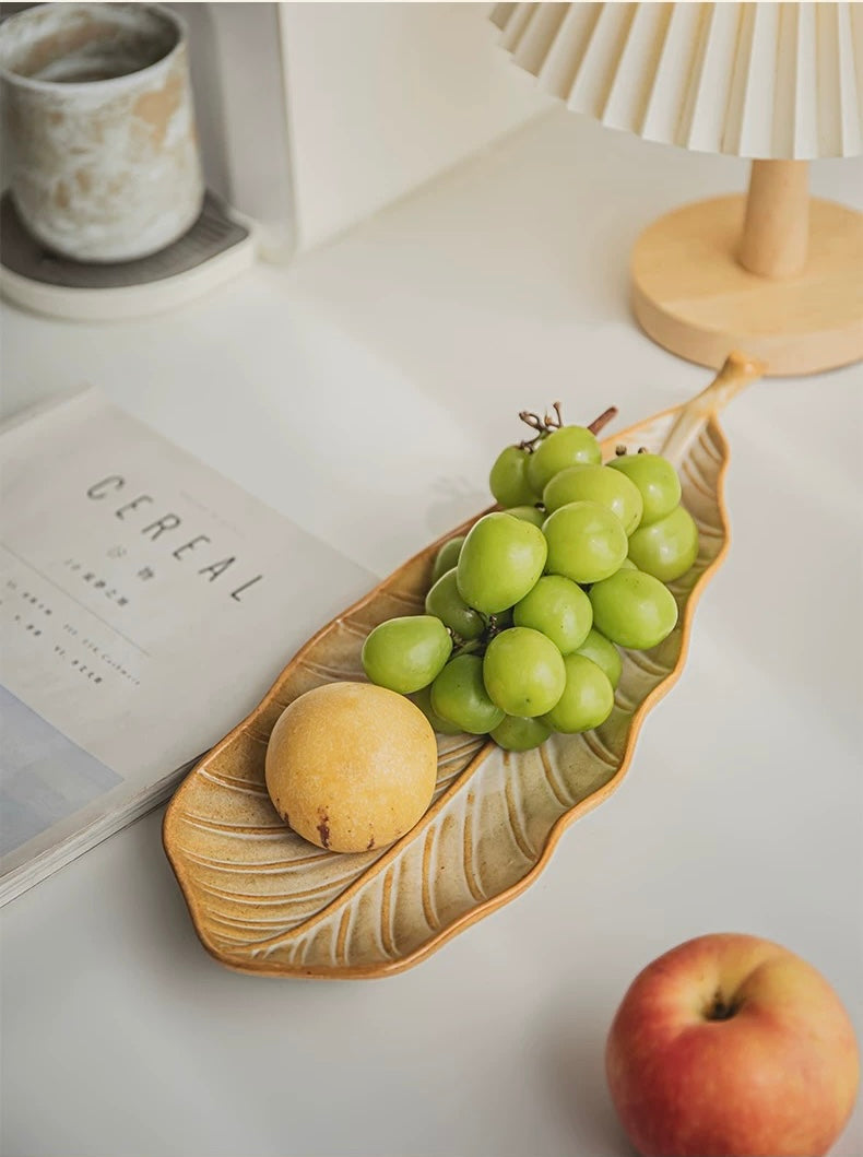 Leaf-shaped ceramic plate with grapes and a peach on a white surface.