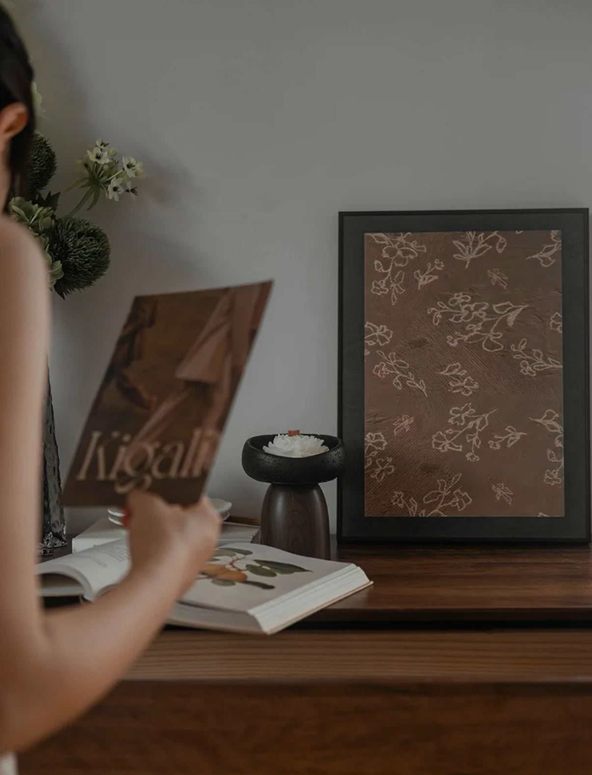 Person holding a book titled 'Kigali' with a framed picture of a floral design on a wooden surface.