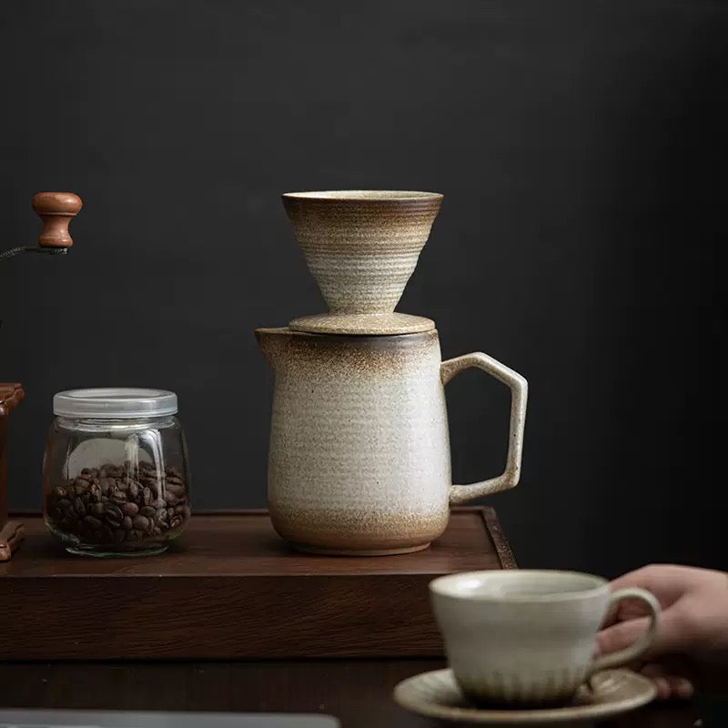 Ceramic coffee maker with a glass of coffee beans and a cup on a wooden surface.