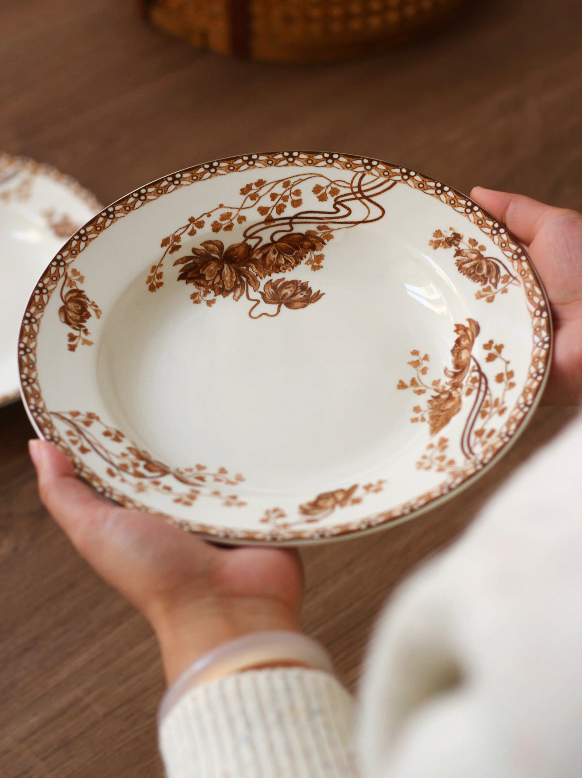 Person holding a decorative ceramic plate with brown floral patterns on a wooden surface