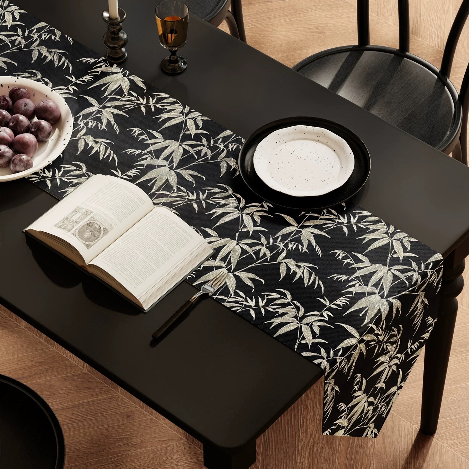 Dining table with a black and white leaf-patterned table runner, open book, and bowl of fruit.