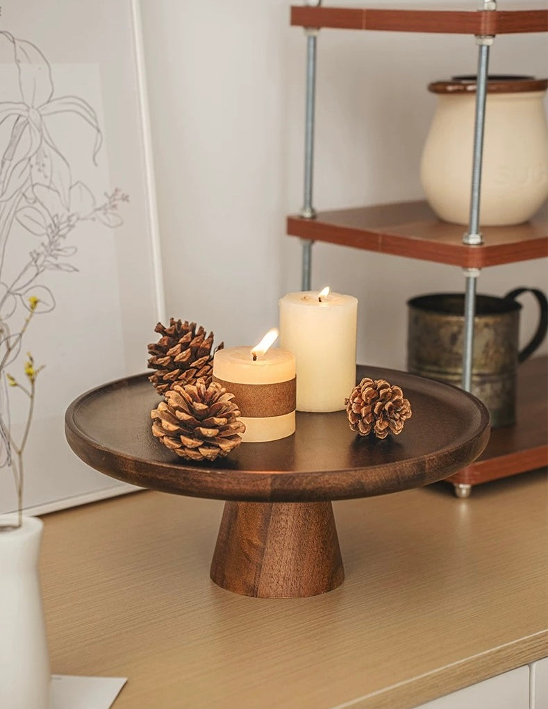 Wooden cake stand with candles and pinecones on a surface, with a shelf in the background.