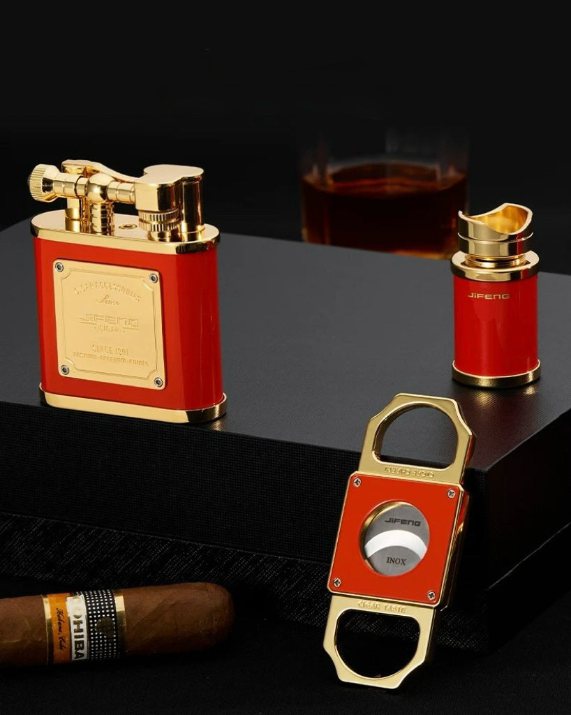 Red and gold lighter, bottle opener, and cigar on a dark surface with a glass of whiskey in the background.