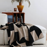 Black and beige patterned blanket draped over a white sofa with a wooden side table and plant in the background.