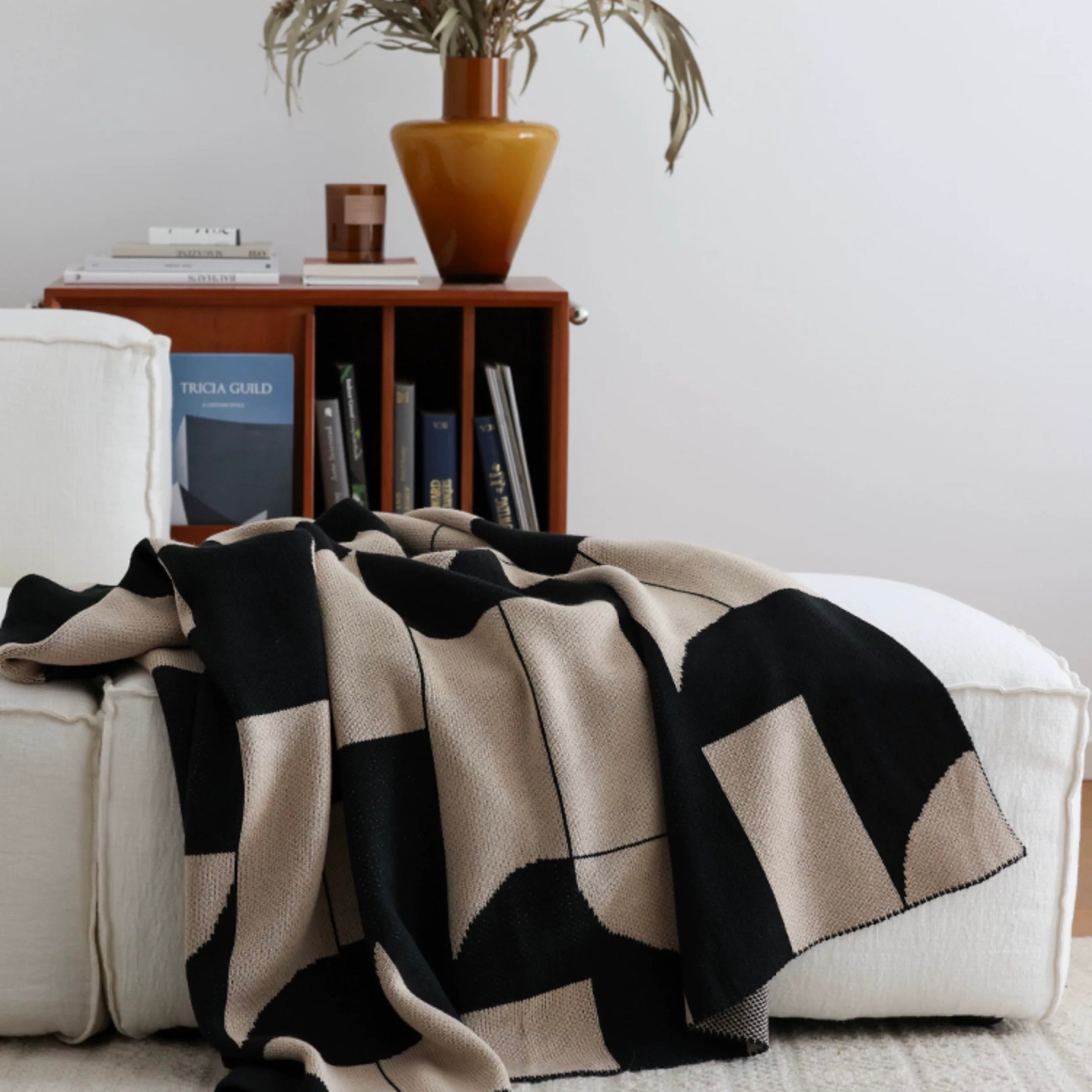 Black and beige patterned blanket draped over a white sofa with a wooden side table and plant in the background.