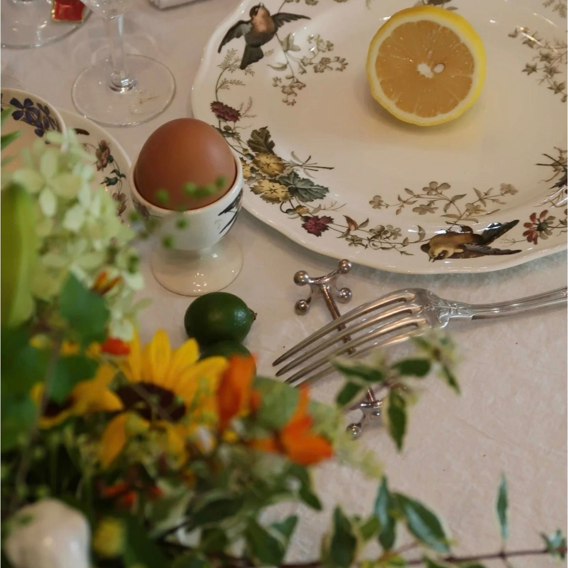 Table setting with floral arrangement, lemon, egg, and silverware on a decorative plate.