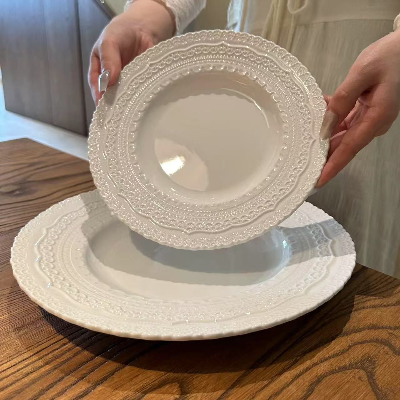 Two white decorative plates stacked on a wooden surface with hands holding them.