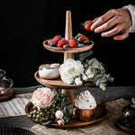 Three-tiered wooden dessert stand with strawberries, flowers, and a cupcake on a dark surface.