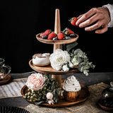 Three-tiered wooden dessert stand with strawberries, flowers, and a cupcake on a dark surface.