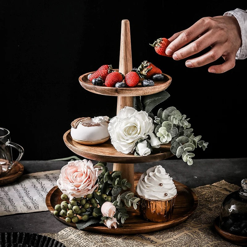 Three-tiered wooden dessert stand with strawberries, flowers, and a cupcake on a dark surface.