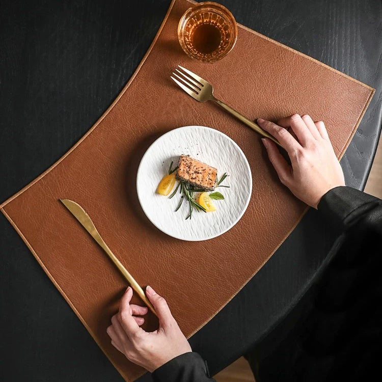 Person setting a table with a plate of food, fork, knife, and glass on a brown placemat.