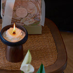 Candle in a wooden holder with a decorative box in the background on a woven mat.