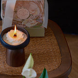 Candle in a wooden holder with a decorative box in the background on a woven mat.