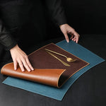 Person unrolling a brown leather placemat with cutlery on a dark surface