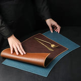 Person unrolling a brown leather placemat with cutlery on a dark surface