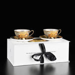Two ornate teacups and saucers in a white box with a black ribbon on a black background