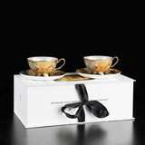Two ornate teacups and saucers in a white box with a black ribbon on a black background
