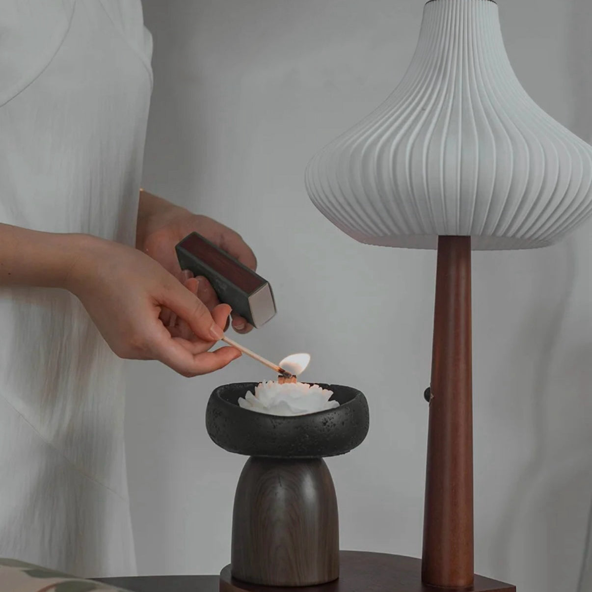 Person lighting a candle in a wooden holder with a modern lamp in the background
