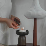 Person lighting a candle in a wooden holder with a modern lamp in the background