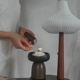 Person lighting a candle in a wooden holder with a modern lamp in the background