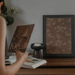 Person holding a book titled 'Kigali' with a framed picture of a floral design on a wooden surface.