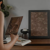 Person holding a book titled 'Kigali' with a framed picture of a floral design on a wooden surface.