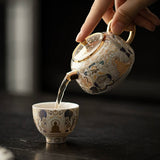 Tea being poured from an ornate teapot into a matching cup on a dark surface.