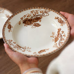 Person holding a decorative ceramic plate with brown floral patterns on a wooden surface