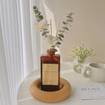 A bottle of 'Floral Petals' diffuser with flowers on a wooden stand on a white surface.