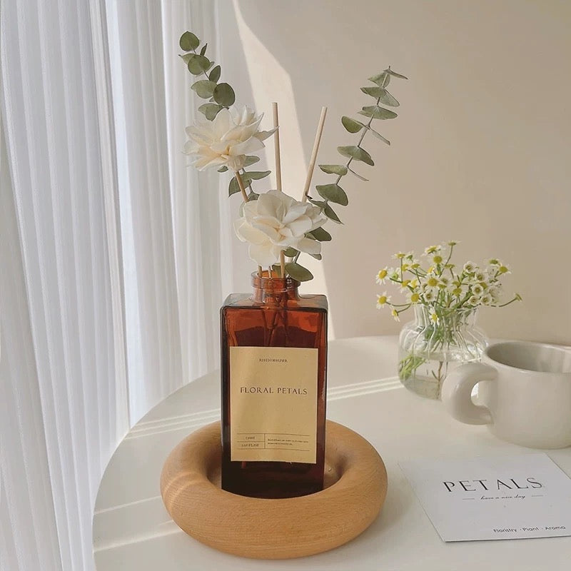 A bottle of 'Floral Petals' diffuser with flowers on a wooden stand on a white surface.
