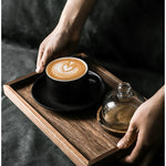 Person holding a wooden tray with a cup of coffee and a glass container on a dark surface
