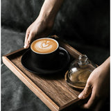 Person holding a wooden tray with a cup of coffee and a glass container on a dark surface