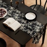 Dining table with a black and white leaf-patterned table runner, open book, and bowl of fruit.