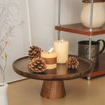 Wooden cake stand with candles and pinecones on a surface, with a shelf in the background.