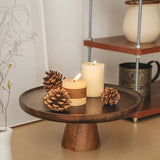 Wooden cake stand with candles and pinecones on a surface, with a shelf in the background.